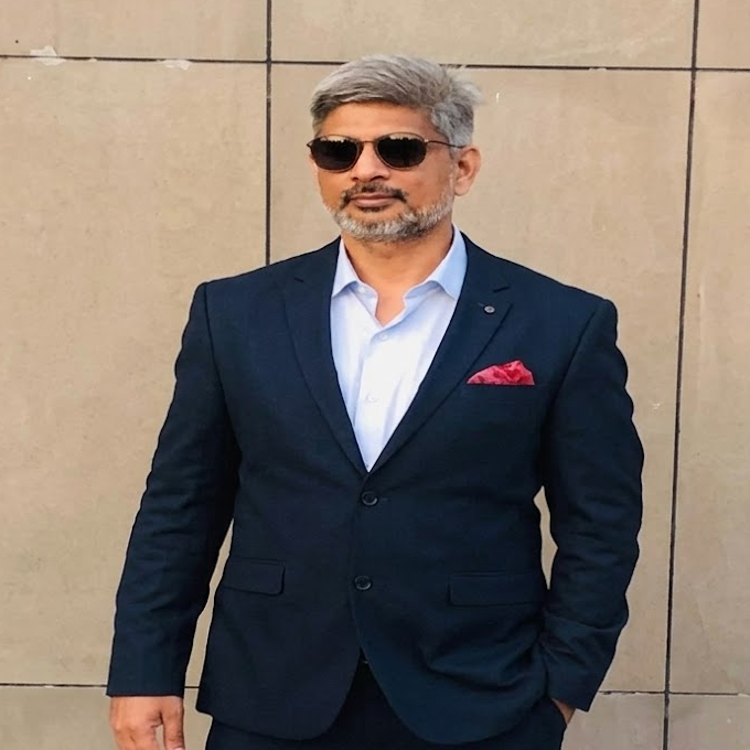 Man in a navy suit with white shirt and red pocket square, wearing sunglasses, posing against a tiled wall.